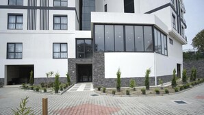 A modern building with a large glass entrance, a stone facade, and a paved courtyard with small trees. - Stylish 1BR Flat Pool View & 10 min Walk to Beach (Avsallar)