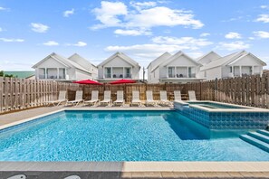 A swimming pool with lounge chairs and umbrellas, surrounded by a wooden fence and white buildings.