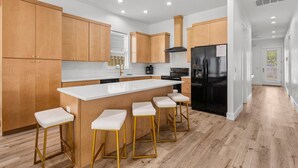 A modern kitchen with wooden cabinets, a large island with bar stools, a black refrigerator, and a window with blinds. - Cobian Pearl with LSV and Game Rooms (Miramar Beach)