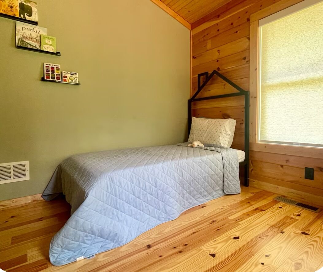 Une petite pièce agréable et chaleureuse, avec un lit, un mur en bois, une fenêtre et une étagère contenant des livres et une boîte.