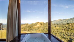 A room with a large window offering a view of rolling hills and a clear sky. - Cabana rústica com jacuzzi VST003 (Anitápolis)