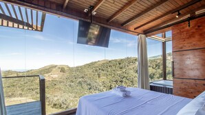 Un dormitorio con un ventanal grande que da a un paisaje montañoso, un televisor de pantalla plana montado en el techo y una cama con sábanas blancas.