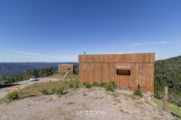 A modern wooden cabin with a large door and a small window, surrounded by a gravel driveway and a sign reading "GARAGEM 43".