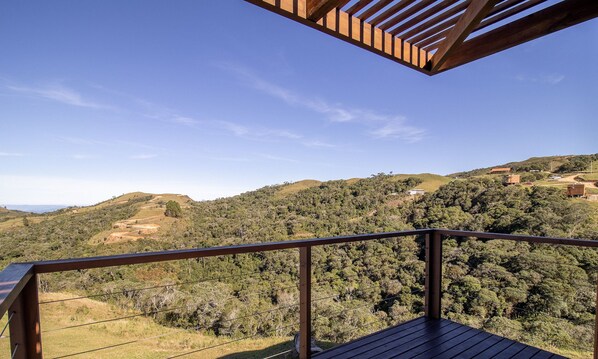 Un balcon en bois, doté d’une rambarde en métal, surplombe un paysage vallonné parsemé de bâtiments.