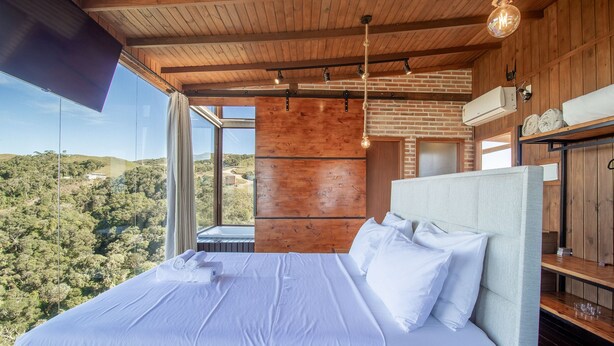A bedroom with a large bed, wooden ceiling, and a view of a forested landscape. - Cabana rústica com jacuzzi VST003 (Anitápolis)