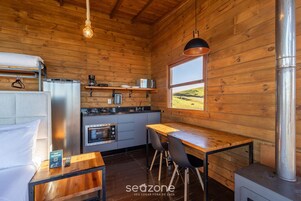 A cozy cabin interior with a wooden interior, a dining area, a kitchen with appliances, and a large window offering a view of the outdoors.