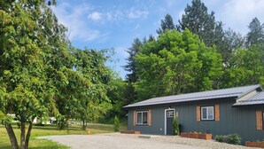 A small, dark-colored building with a gravel driveway, surrounded by lush green trees and a clear sky.