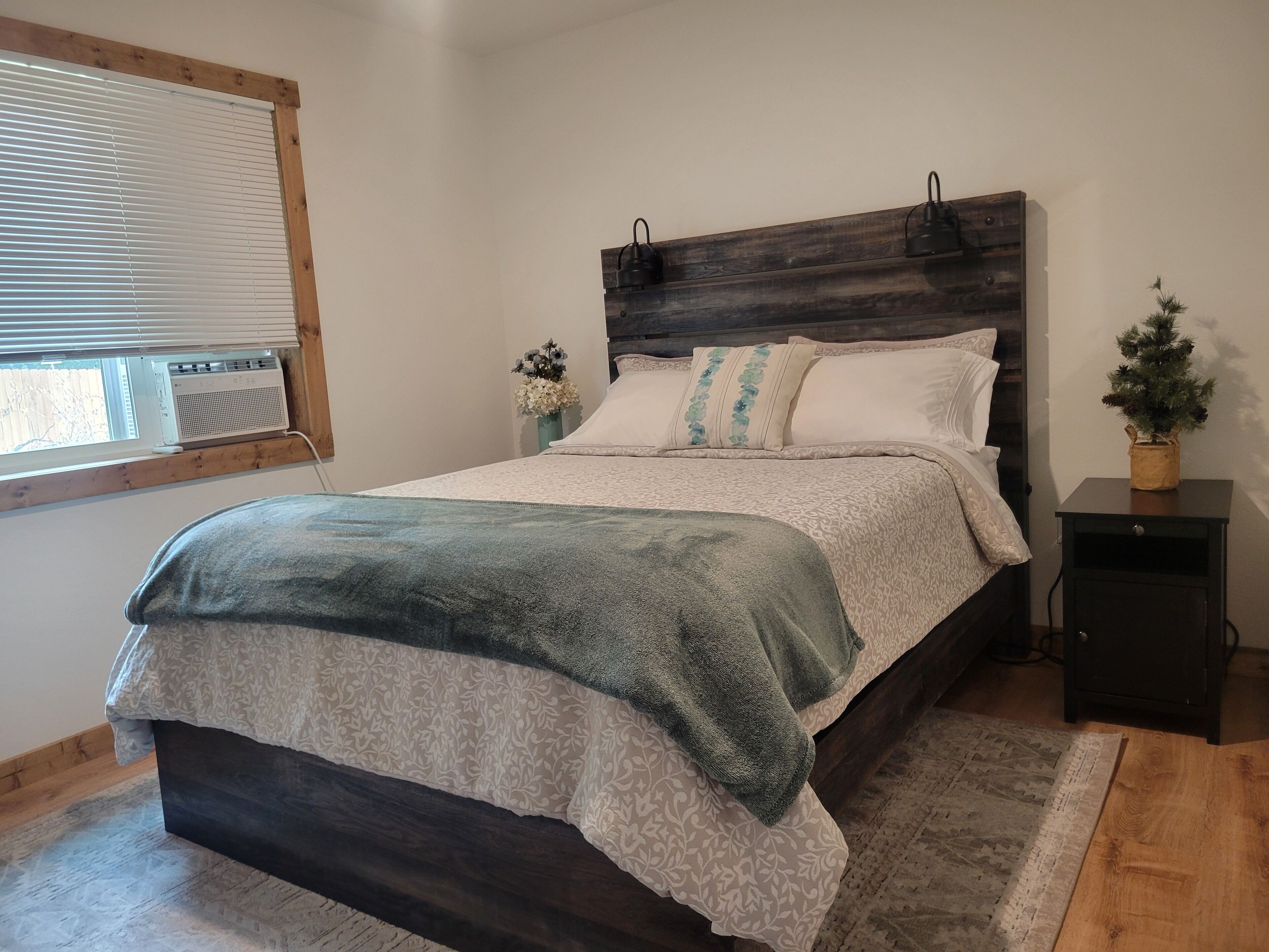 Une chambre à coucher comprenant un lit, une table de chevet, une fenêtre avec des stores, un radiateur et une petite plante en pot.