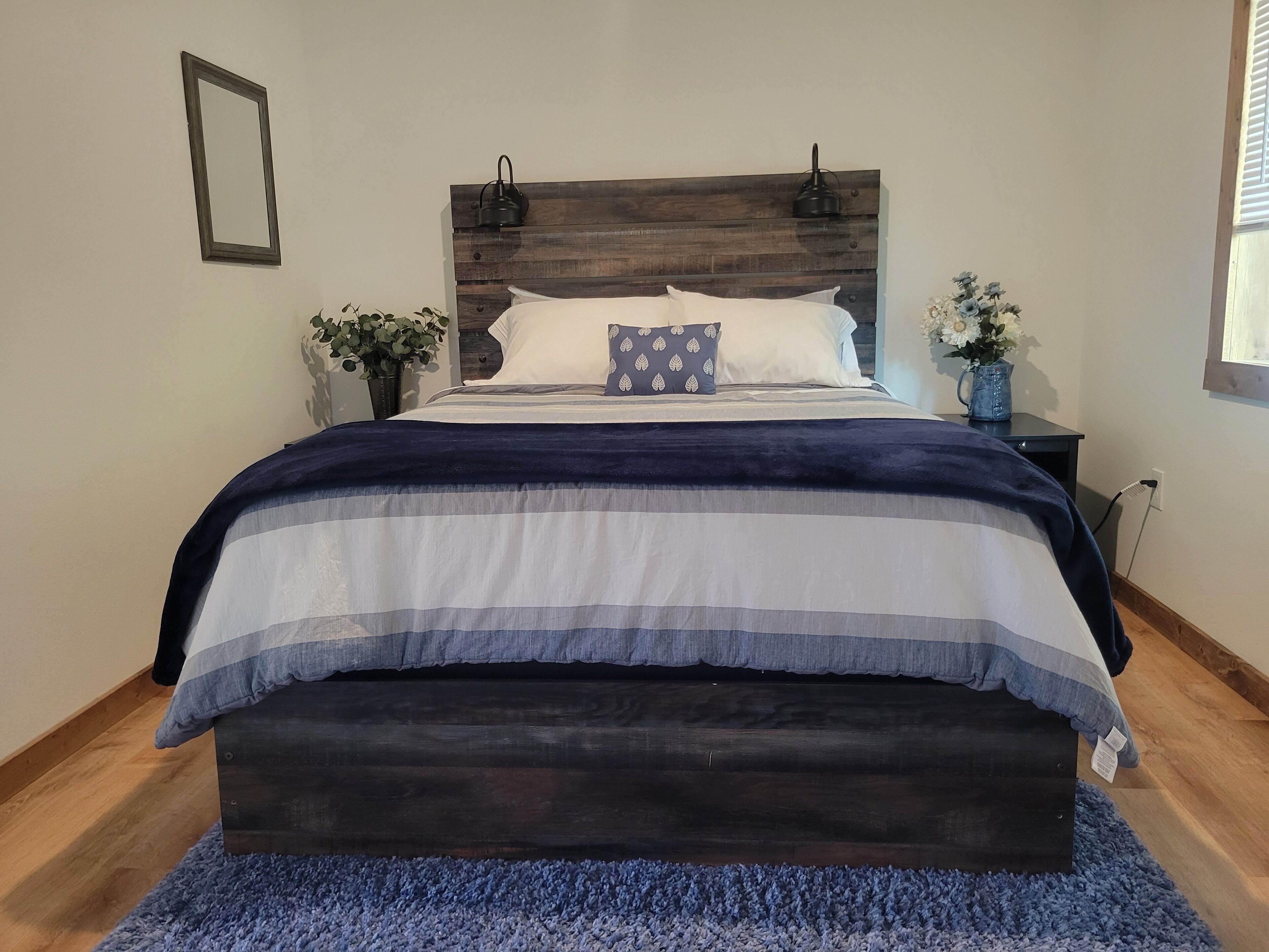 A bed with a dark wooden headboard, white and blue striped bedding, and a blue rug. There are two potted plants, one on each side of the bed.