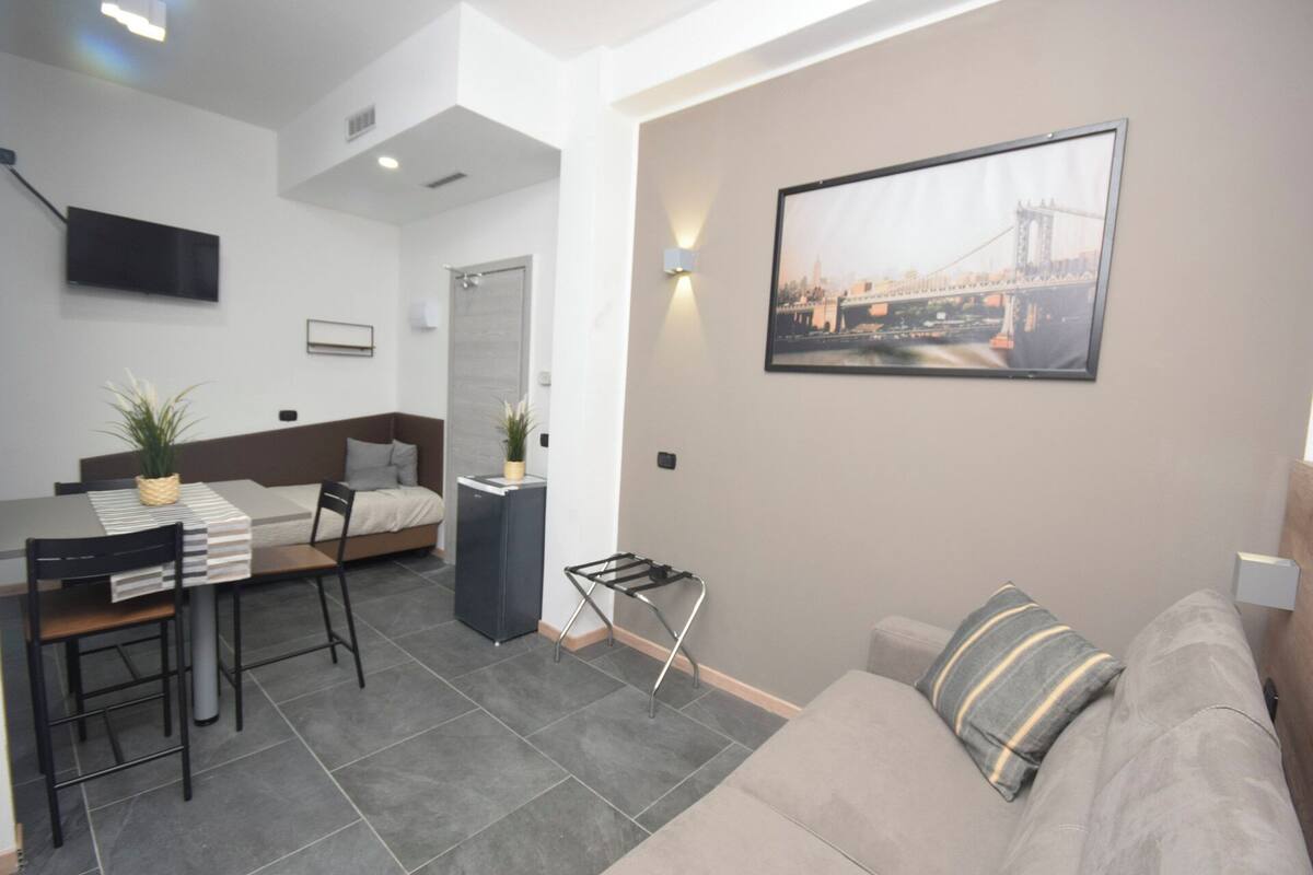 A modern living room with a sofa, a dining area with a table and chairs, a bed, and a TV mounted on the wall.