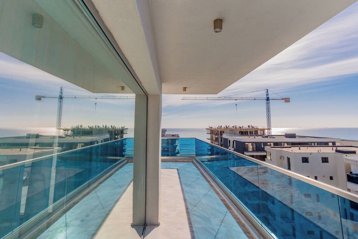 A modern rooftop with glass railings, a pool, and a view of the sea and construction cranes.