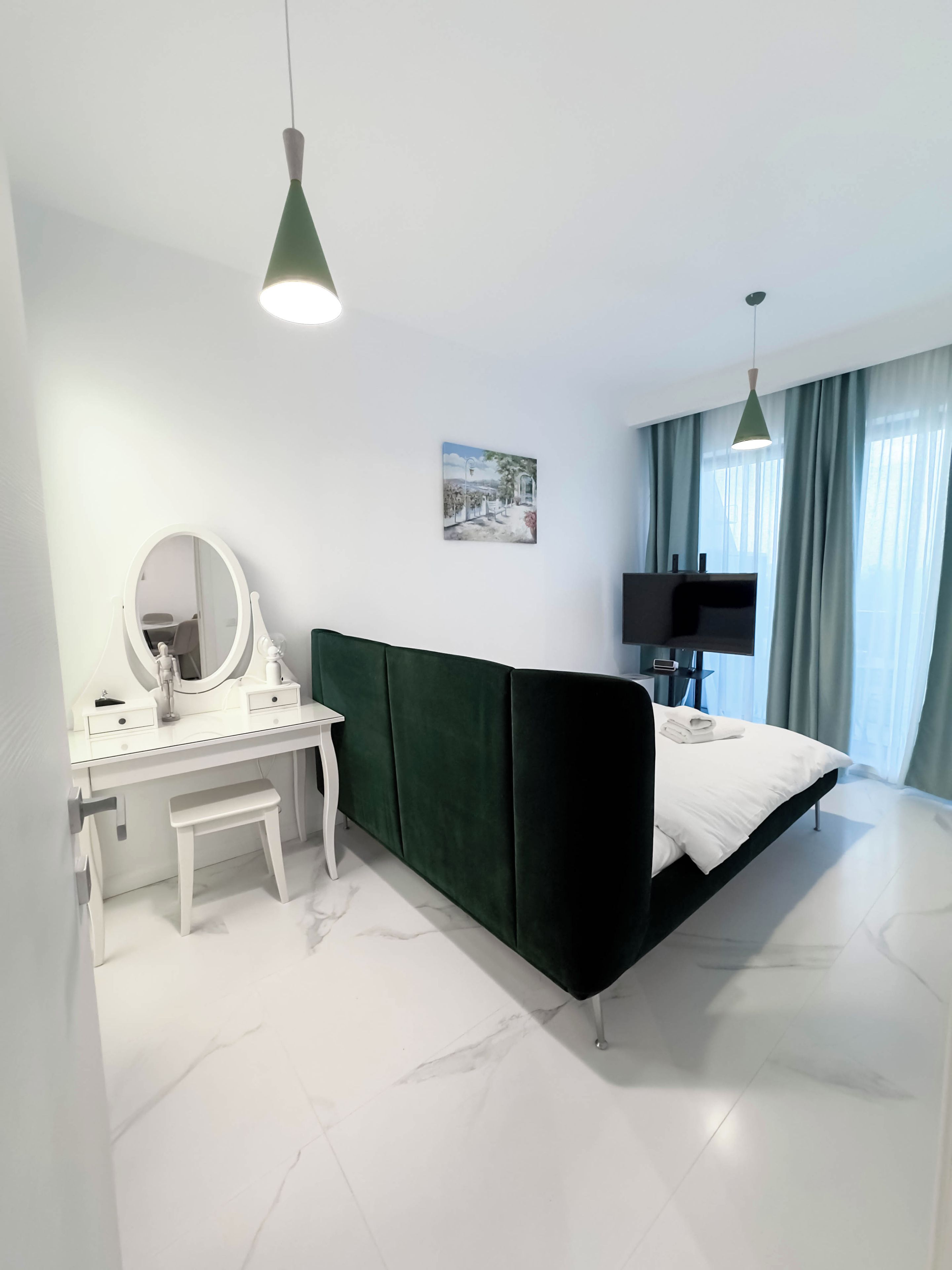 A modern bedroom with a white vanity, a green upholstered bed, a flat-screen TV, and a painting on the wall.