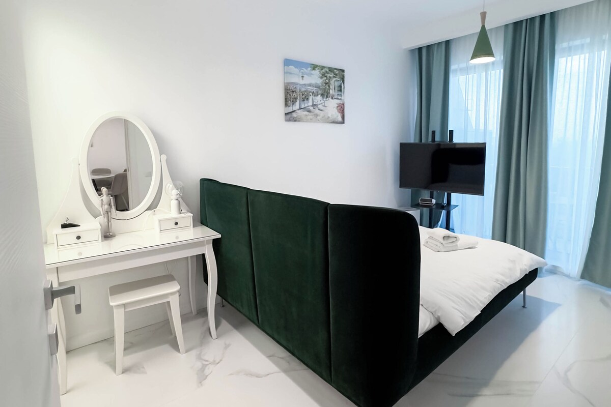 Une chambre moderne avec une coiffeuse blanche, un lit recouvert d’un tissu vert, un téléviseur à écran plat et un tableau accroché au mur.