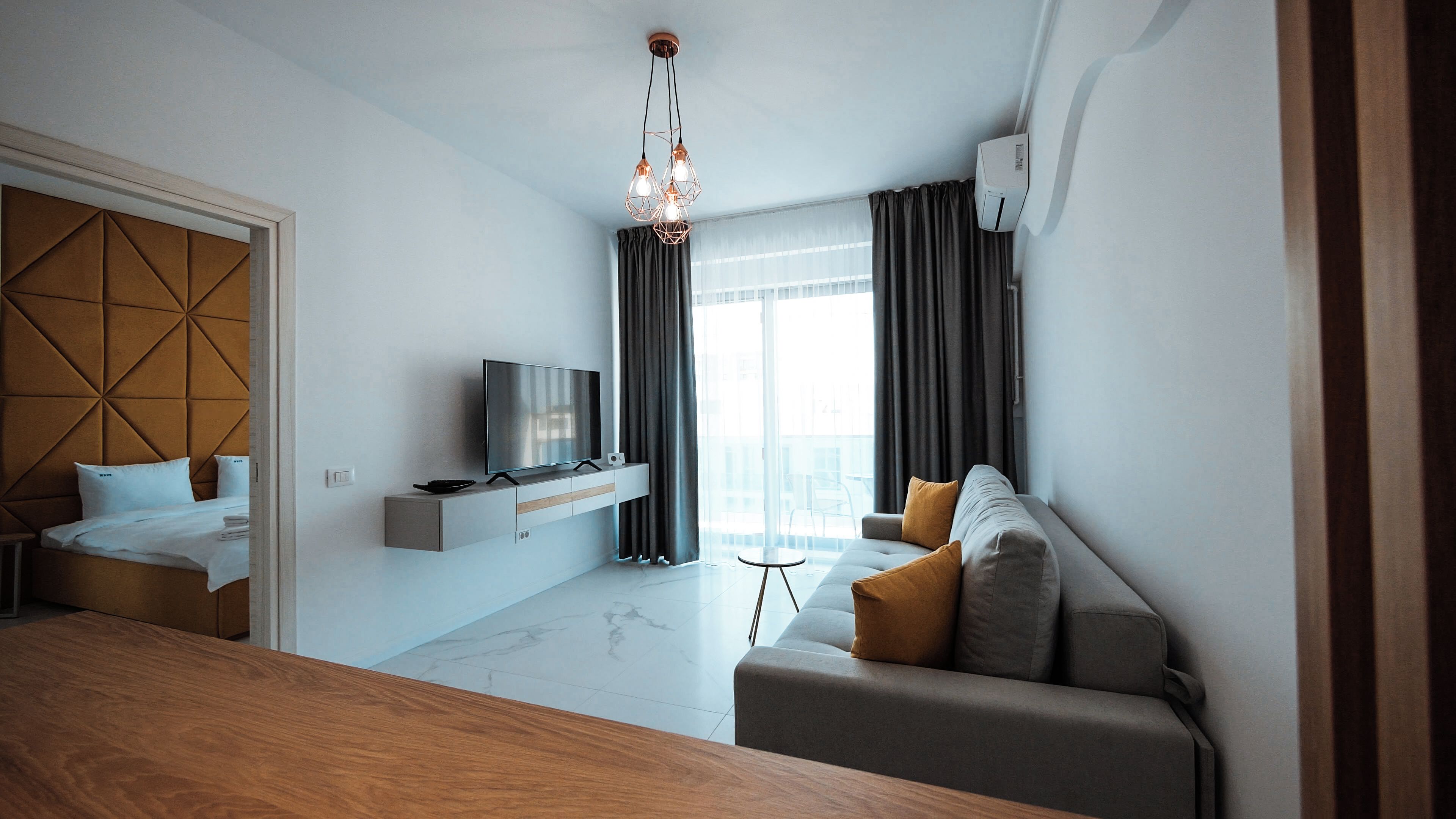 A modern living room with a sofa, a TV on a built-in shelf, a dining table, and a large window with curtains.