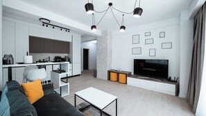 A modern living room with a grey sofa, a coffee table, a TV, and a kitchen area with white cabinets and a stone accent wall. - Wave 4 Apartments (Navodari)