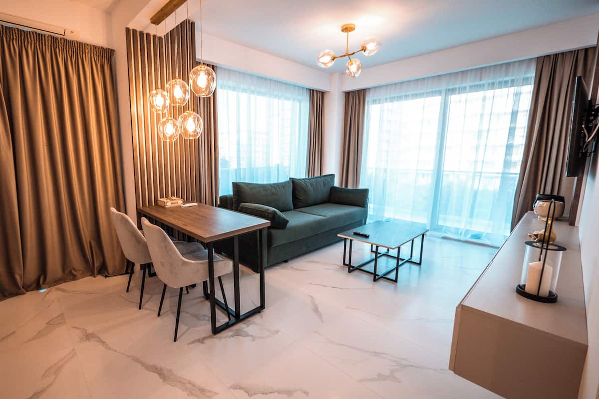 A modern living room with a sofa, a dining table, and chairs. There is a chandelier, a television, and a large window with curtains.