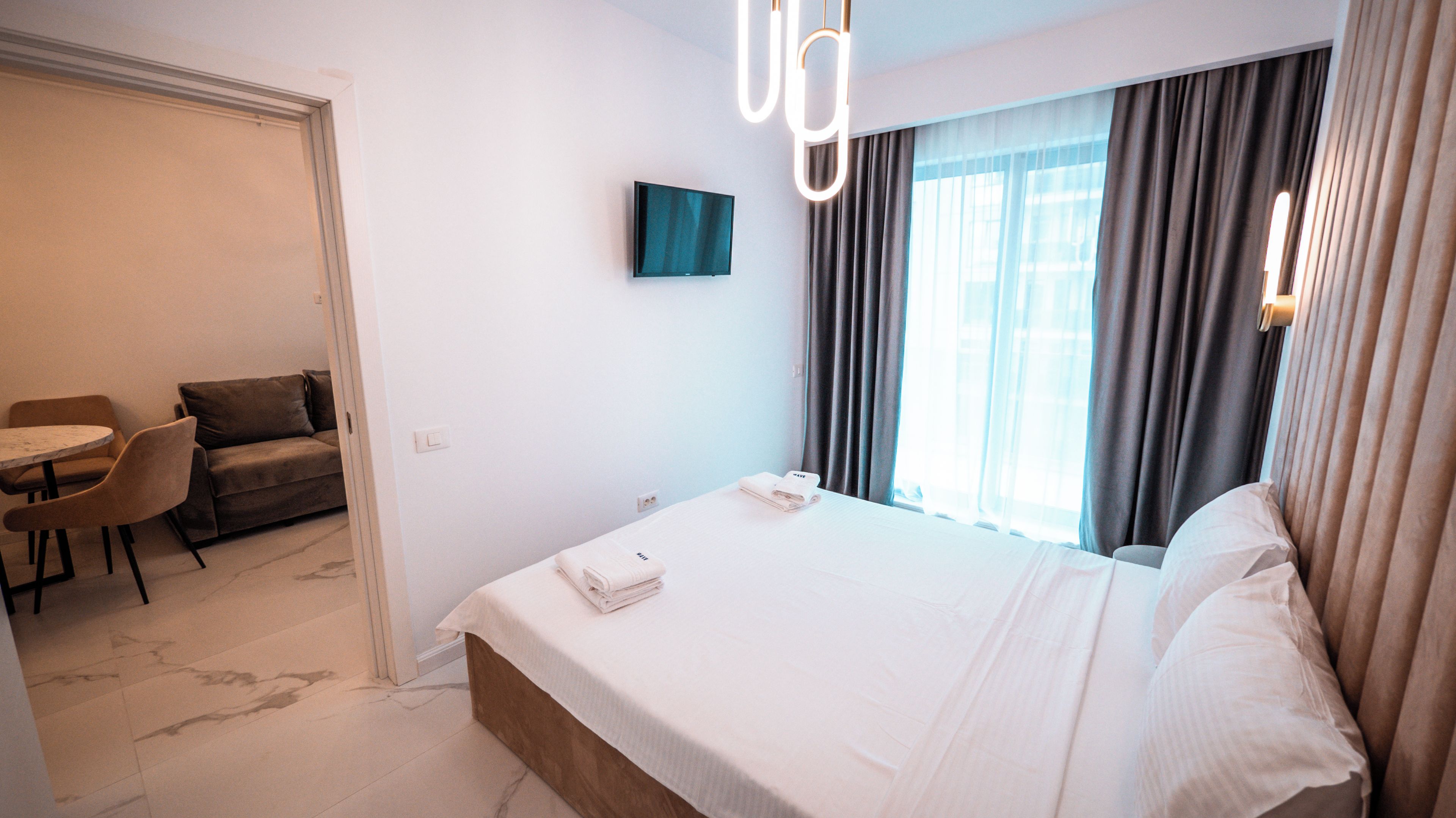 A hotel room with a bed, a TV, a dining area with a table and chairs, and a window with curtains.