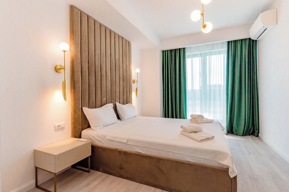 A modern hotel room with a wooden headboard, a bedside table with a lamp, a neatly made bed, and a large window with green curtains.