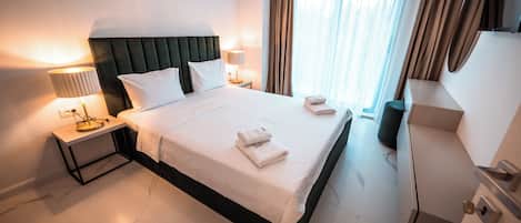 Habitación de hotel con una cama grande, dos mesitas de noche con lámparas, un aparador y una ventana con cortinas.