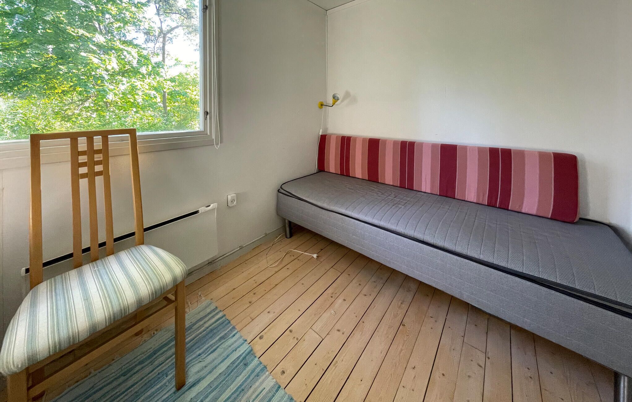 Ein Zimmer mit Holzboden, einem Streifenstuhl, einem grauen Sofa mit einem rot-pink gestreiften Kissen und einem Fenster mit Blick auf Bäume.