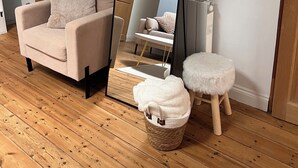 Una habitación con piso de madera, un sofá color beige, un espejo grande, un taburete pequeño y una cesta.