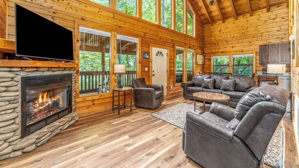 A log cabin interior with a stone fireplace, leather sofas, and large windows offering a view of the forest. - Mountain Falls 14 | Family Friendly + Hot Tub (Sevierville)