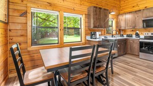 A wooden cabin interior with a dining table, chairs, a kitchen with wooden cabinets, and large windows. - Mountain Falls 14 | Family Friendly + Hot Tub (Sevierville)