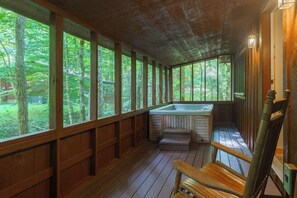 Una terraza de madera con un jacuzzi, bancos de madera y ventanas amplias que ofrecen vistas del bosque circundante.