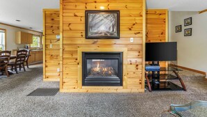 Una sala de estar con chimenea, paredes de paneles de madera, un cuadro enmarcado sobre la chimenea, un televisor en un pedestal y un comedor al fondo.