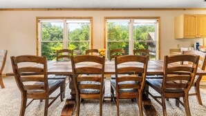 Un comedor con una mesa y sillas de madera, ventanas amplias con vista a follaje, y una zona de cocina con armarios de madera.