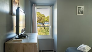 Une pièce avec un bureau, une fenêtre donnant sur des arbres et un parapluie bleu, un lit avec une serviette blanche pliée, et un tableau encadré au mur.