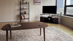 Een woonkamer met een donkerhouten salontafel, een boekenkast, een flatscreen-tv op een houten standaard en een groot raam met uitzicht over de stad.