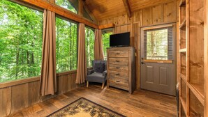 A wooden cabin interior with large windows, a chair, a dresser, and a television. - Little Bit of Heaven | Secluded + Woodland Haven (Sevierville)