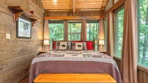 A wooden cabin bedroom with a bed, bench, and large windows overlooking a forest. - Little Bit of Heaven | Secluded + Woodland Haven (Sevierville)