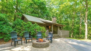 A lodge with a stone fire pit surrounded by Adirondack chairs, set in a forested area with a wooden deck and a porch. - Little Bit of Heaven | Secluded + Woodland Haven (Sevierville)