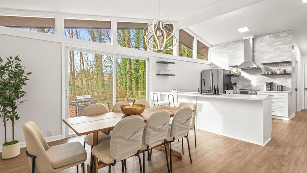 A modern kitchen with a large island, white cabinetry, and a view of trees through large windows.