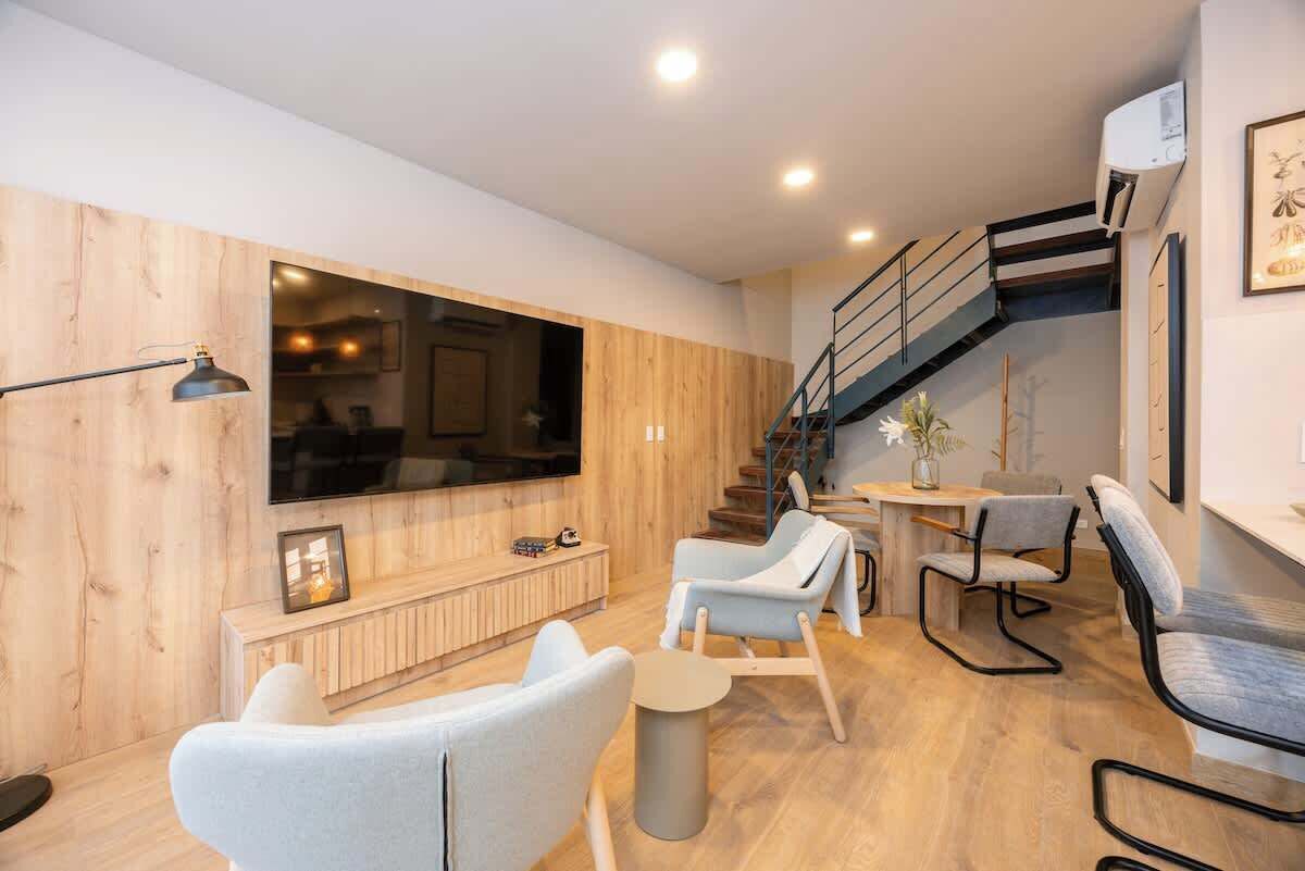 A modern living room with a large flat-screen TV, wooden panel walls, a staircase, and a dining area with light-colored chairs.
