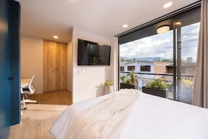 A modern bedroom with a large bed, a TV mounted on the wall, a desk with a chair, and a view of the city through large windows.