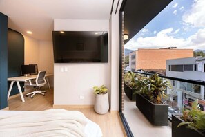 A modern bedroom with a bed, a desk, a chair, a TV, and a balcony with plants.