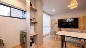 A modern living room with a built-in washer, a dining area with a table and chairs, a large TV mounted on a wooden wall, and a window with a view of a brick building.
