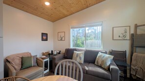 Un salon chaleureux avec un plafond en bois, une grande fenêtre donnant sur les arbres, un canapé marron agrémenté de coussins à motifs, un fauteuil beige et une petite table sur laquelle se trouvent un panier et un objet décoratif.
