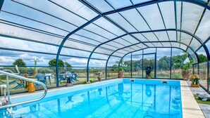 A covered swimming pool with clear glass walls, a diving board, and potted plants. - Charmante Maison avec piscine (Lacropte)