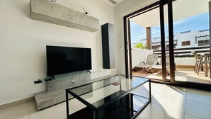 Un salon moderne doté d’un téléviseur à écran plat, d’une table basse en verre et d’une porte-fenêtre coulissante donnant sur une terrasse extérieure avec des chaises.