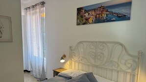 A bedroom with a bed, a nightstand, a lamp, a painting, and a window with curtains. - Pippo a vernazza rooms (Vernazza)