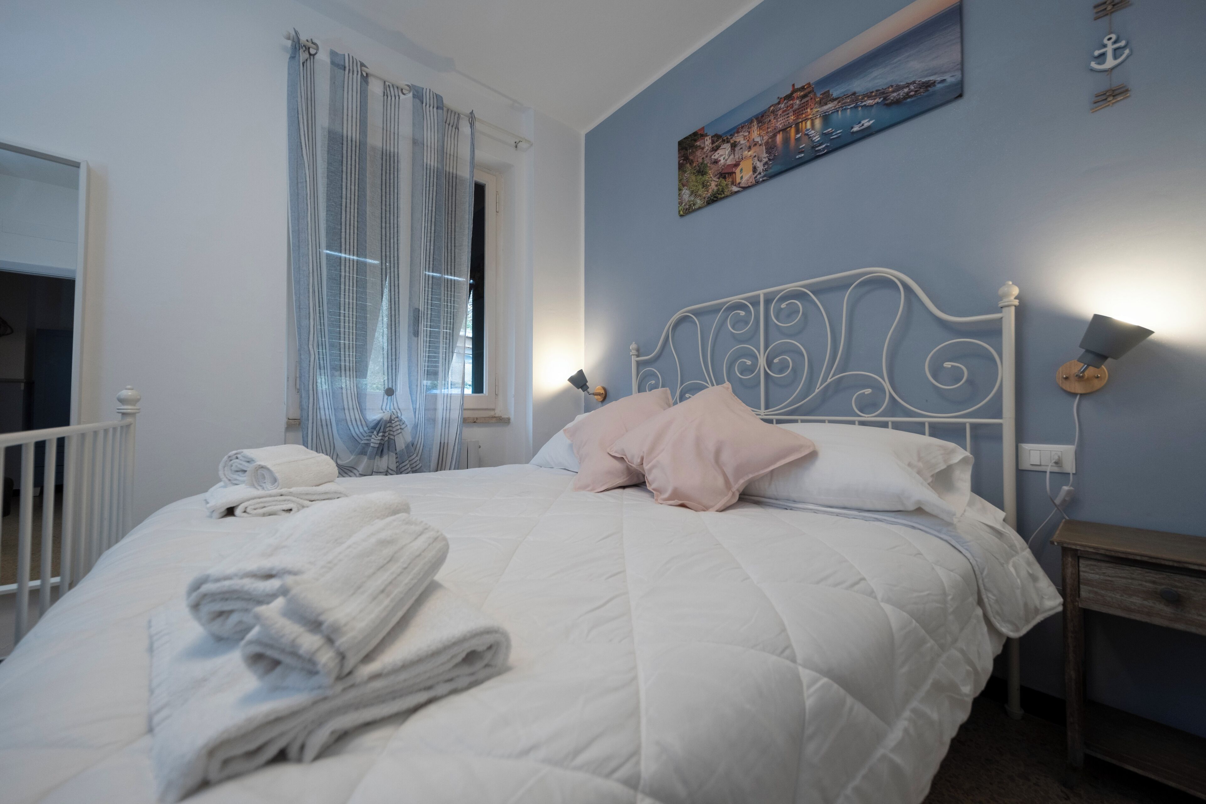 A bedroom with a bed, pillows, a rolled towel, a window with curtains, a wall-mounted lamp, and a painting.