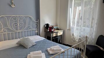 A neatly made bed with a blue quilt, pillows, and folded towels. There is a small bedside table with a kettle and a box. A window with a floral curtain allows natural light into the room.