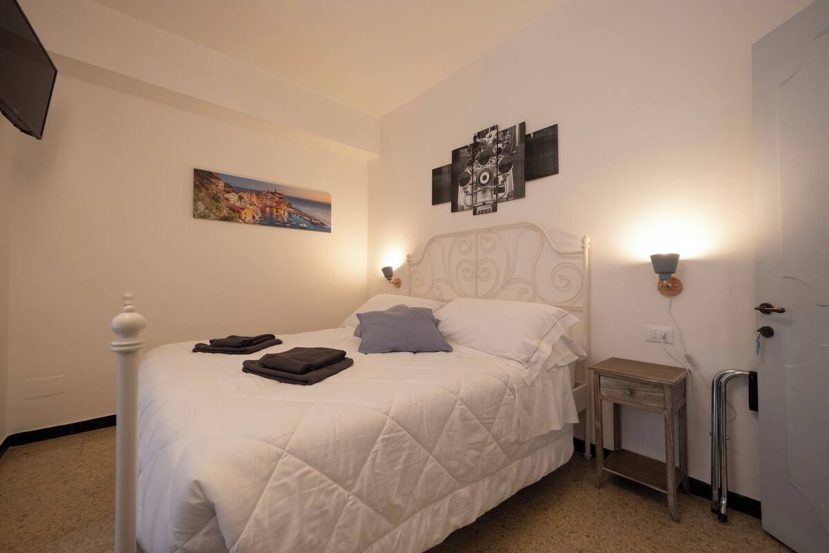 Un lit bien fait, avec des oreillers et une couverture pliée, une table de chevet avec une lampe, une œuvre d’art accrochée au mur et une porte avec une poignée.