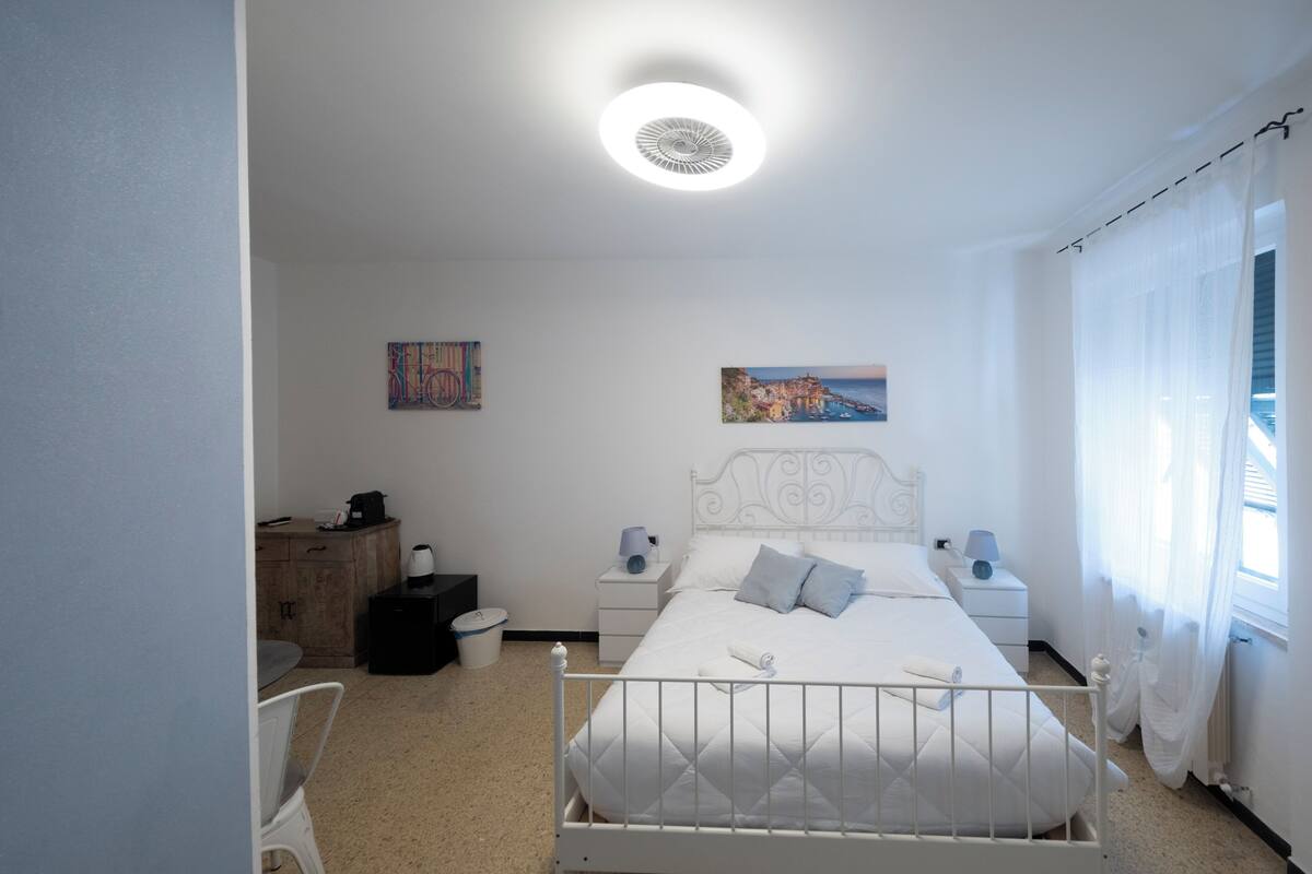 Une chambre à coucher avec un lit blanc, des tables de chevet, une commode, une chaise, un tableau au mur et une fenêtre avec des rideaux.