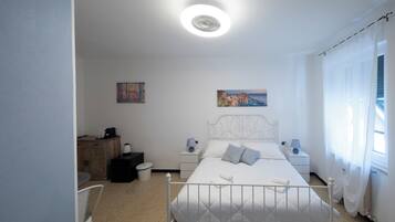 A bedroom with a white bed, bedside tables, a nightstand, a chair, a painting on the wall, and a window with curtains.
