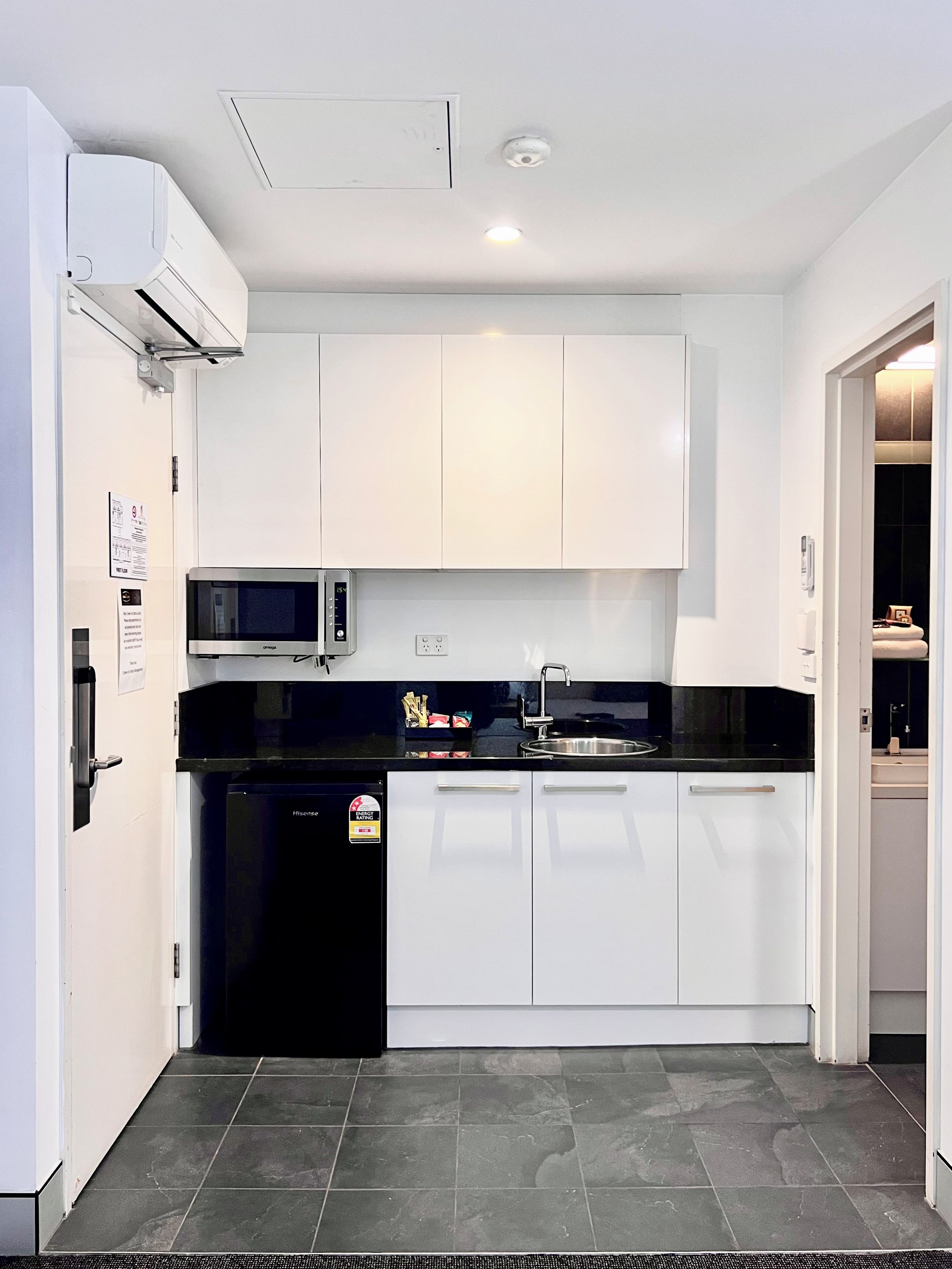 Una cocina compacta con armarios blancos, encimera negra, microondas integrado y refrigerador.